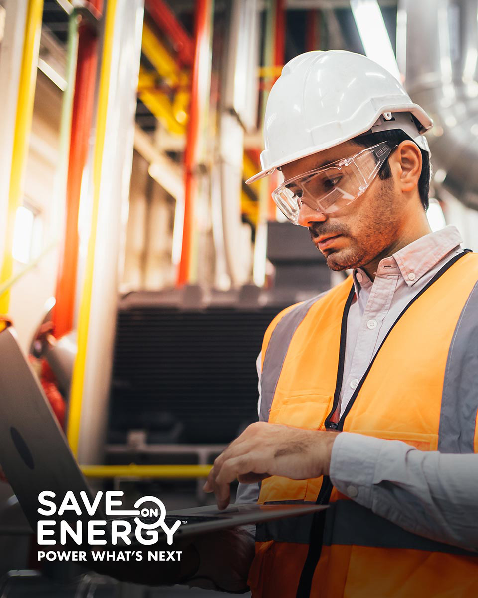 A man in a white hard hat, yellow safety vest and safety glasses types on a computer