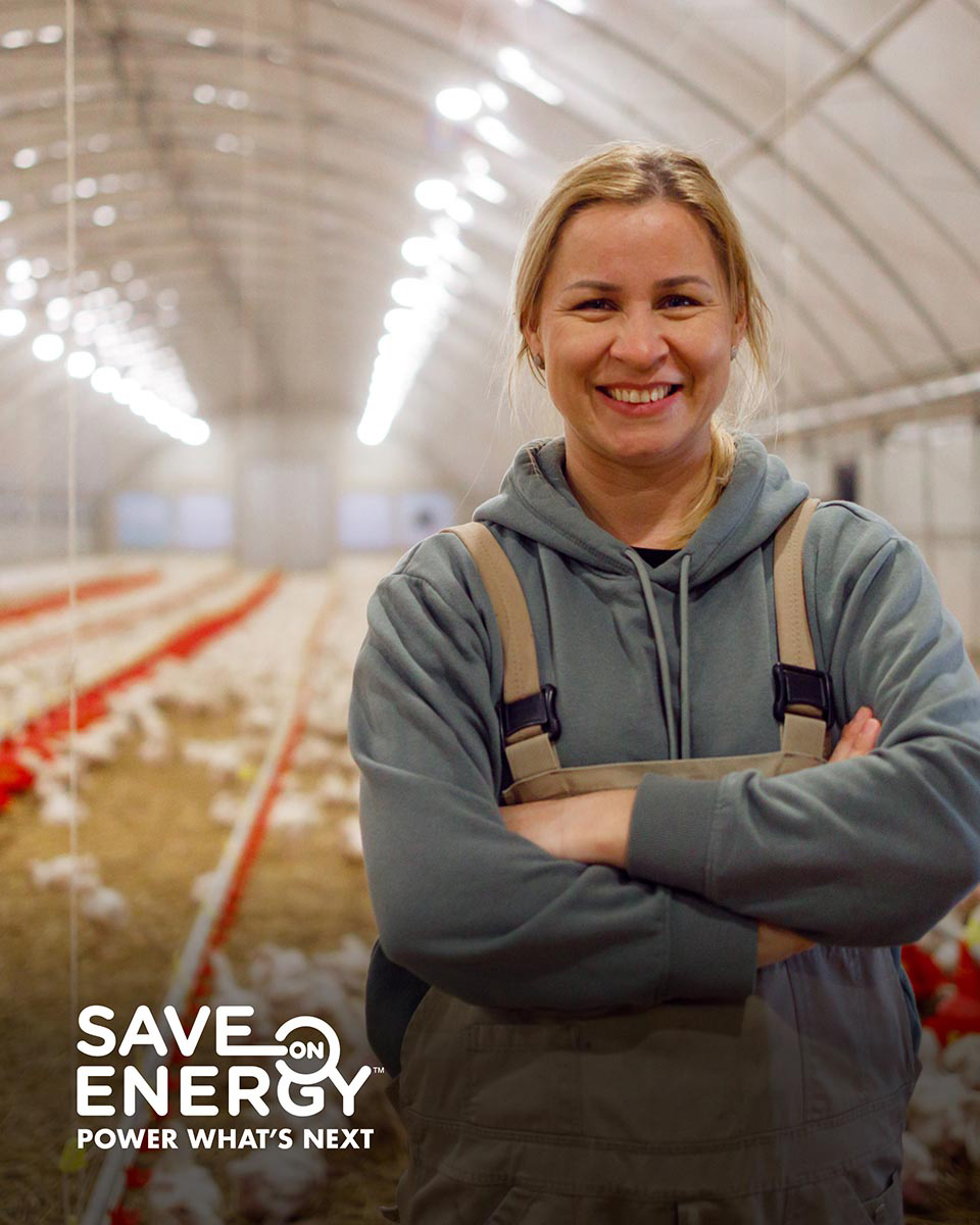 A smiling woman in coveralls stands in front of a barn with chickens
